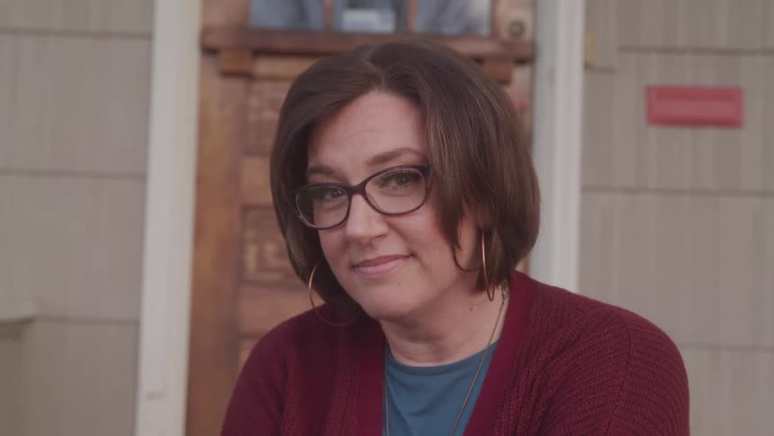 A woman with glasses gives a subtle smile while sitting outside a house