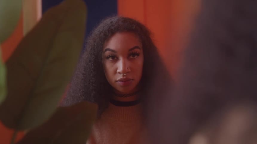 A young woman with curly hair gazes softly into a mirror with a plant in the foreground and an orangeandblue wall behind her