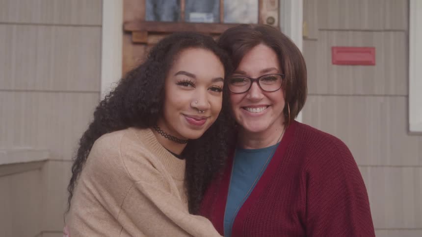 A mother and daughter smile warmly while embracing in front of their home