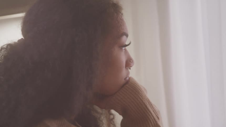 A curlyhaired woman gazes through sheer white curtains with soft sunlight streaming through a window