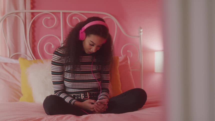 Curly haired woman sitting on bed interacting with smartphone and wearing pink headphones