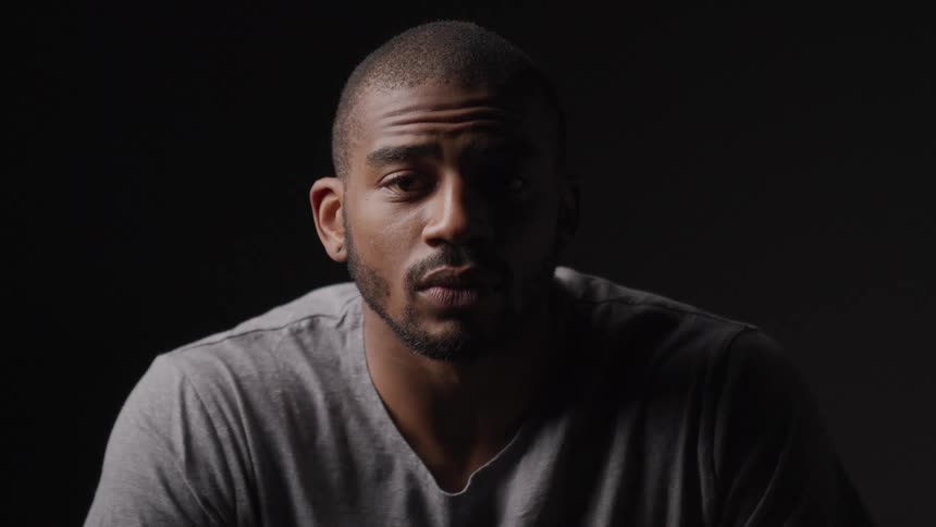 A man sits under dramatic lighting gazing directly at the camera