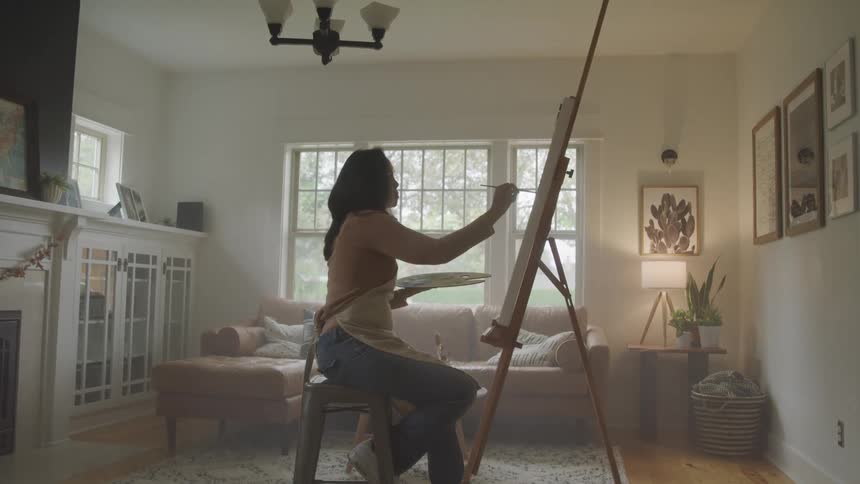 A woman sits in her living room painting on a canvas