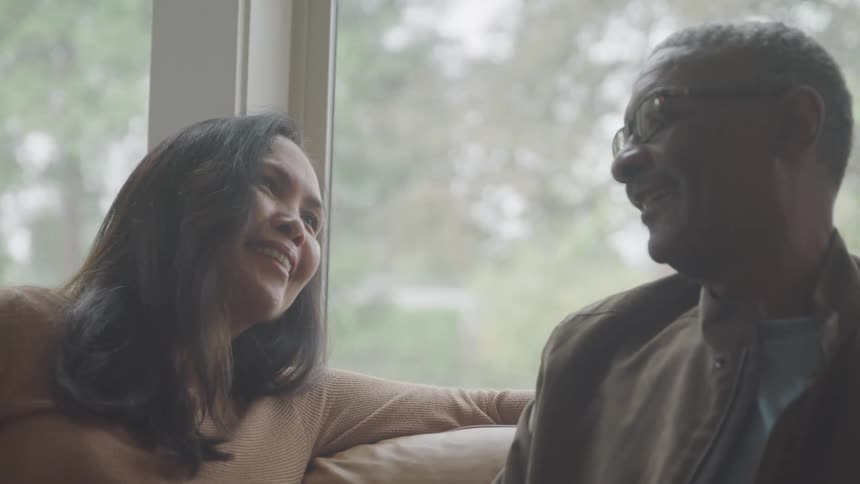 A couple sits by a window smiling warmly at each other