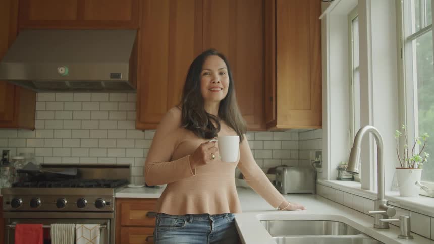 Woman holding a mug with a happy expression while standing in the kitchen