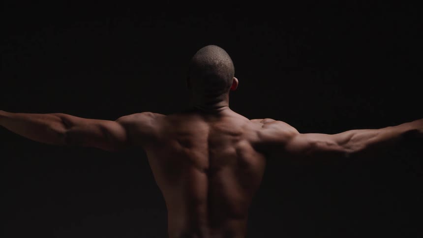 A shirtless black man raises both hands to shoulder level displaying his muscles in a dark room