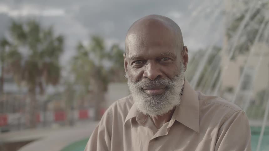 An elderly black man with a bald head and gray facial hair sits beside a fountain in the city around midday