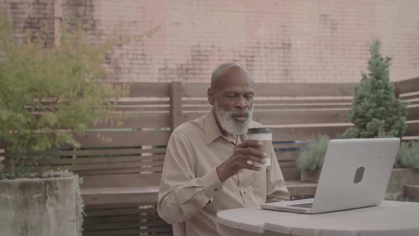 An older man works on his laptop while enjoying coffee at an outdoor table