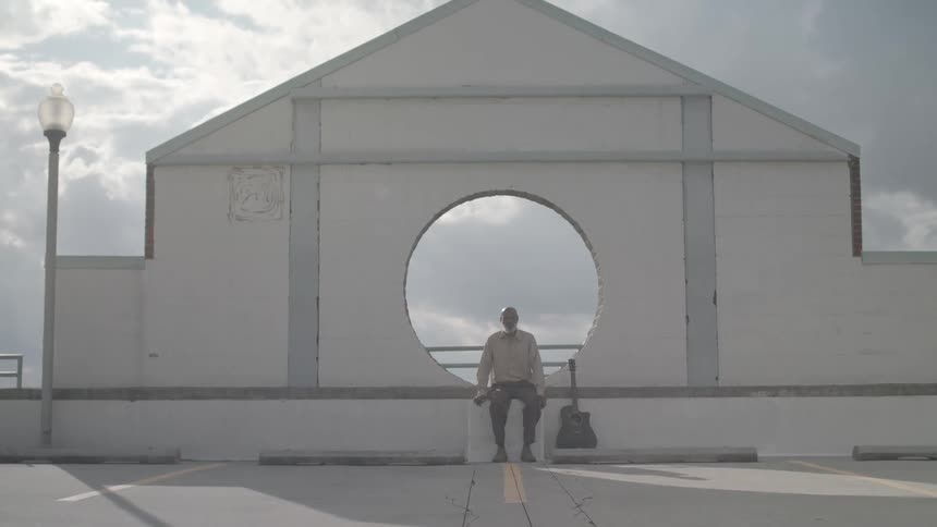 A man sits on a ledge with a guitar next to him framed by a circular opening in a white building under a partly cloudy sky