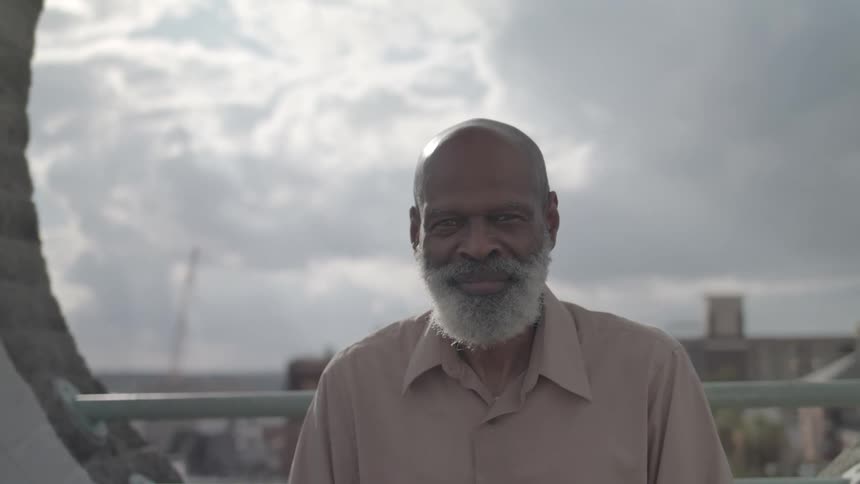 A bald elderly black man with a gray beard sits outdoors around midday