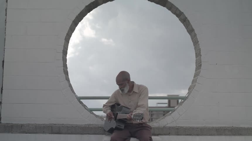 An elderly man sits in a circular wall opening playing a guitar outdoors