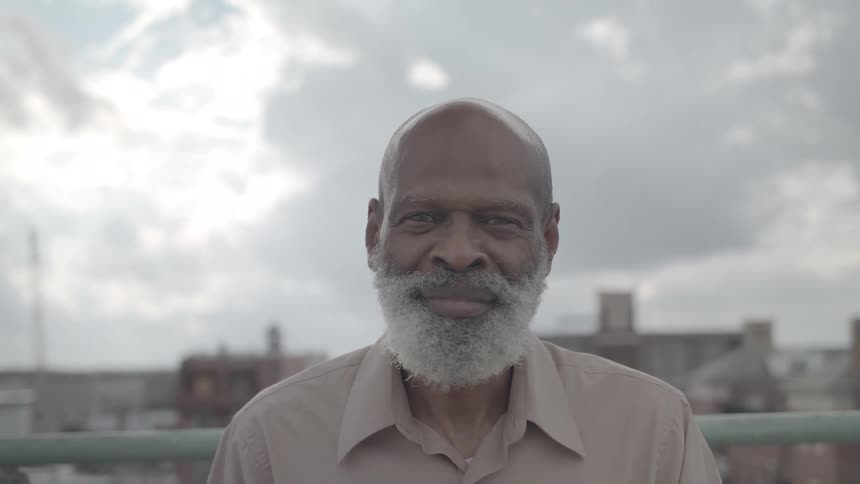 An elderly black man with a bald head and a long gray beard stands outdoors on a cloudy day