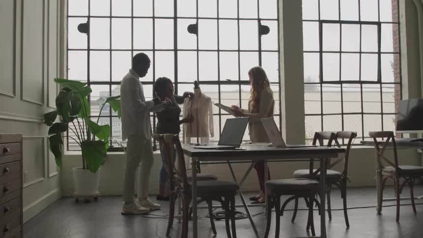 A fashion designer works on a dress design in an office workspace around midday with a man and a woman helping her