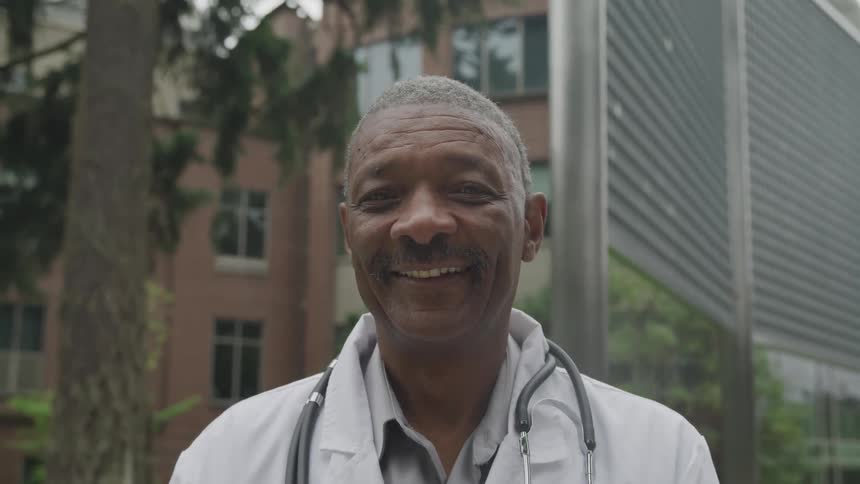 A smiling male doctor stands outdoors with a stethoscope around his neck