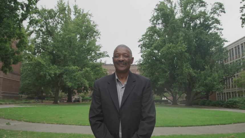 A smiling man in a suit stands confidently on a green campus lawn