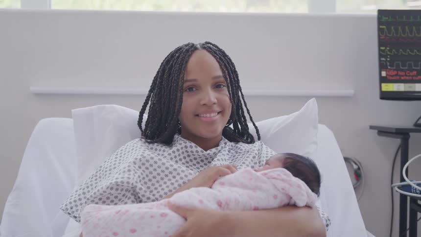 A person in a hospital gown cradles a newborn wrapped in a pink and white blanket with a vital signs monitor in the background