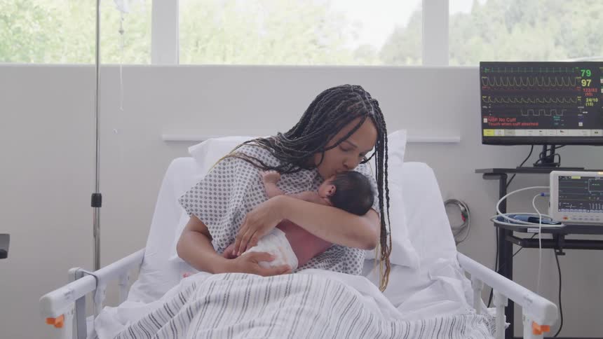 A woman in a hospital bed with a newborn baby in her arms kissing the baby on the head