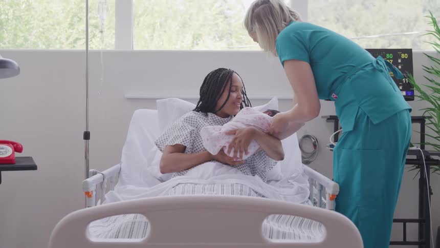 A nurse gently hands a newborn baby to a smiling mother in a hospital bed