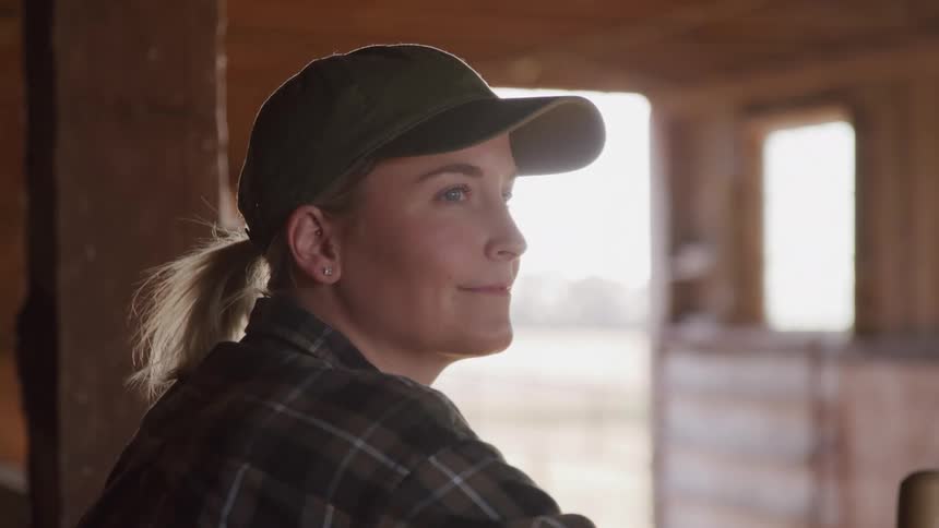 A woman wearing a dark cap and a plaid shirt looks to her right with a soft expression in what appears to be a barn