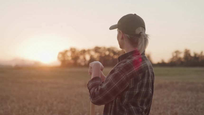 Person in cap stands in field at sunset with shovel