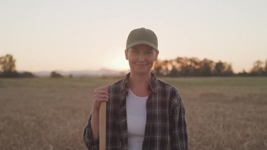 A woman stands in a field at sunset holding a farming tool and smiling at the camera