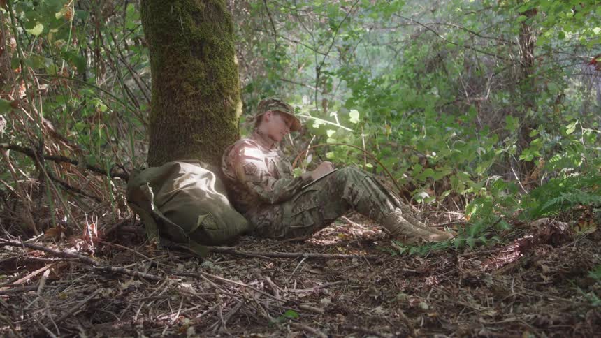 A soldier sits against a tree in a forest writing a letter in a quiet moment