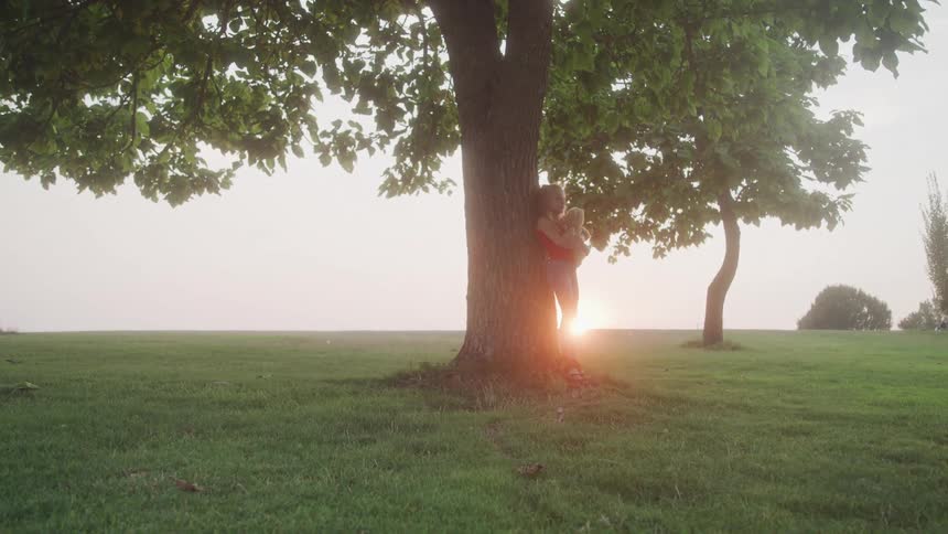 Girl hugs teddy bear while standing under a tree at sunset