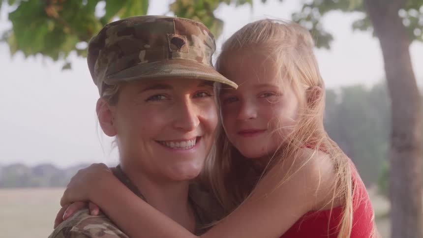 A smiling soldier holds a young girl in a warm embrace outdoors