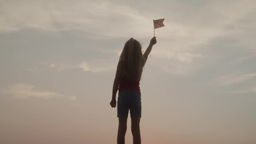 A young child stands outdoors raising a flag into the sky at sunset