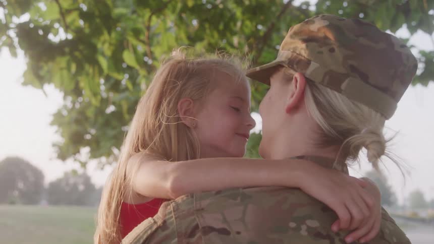 A girl with blonde hair hugs a woman in military uniform under a tree