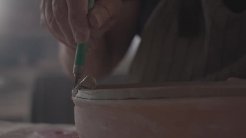 Close up of a man using a bladed tool to carve the edges of pottery in a room