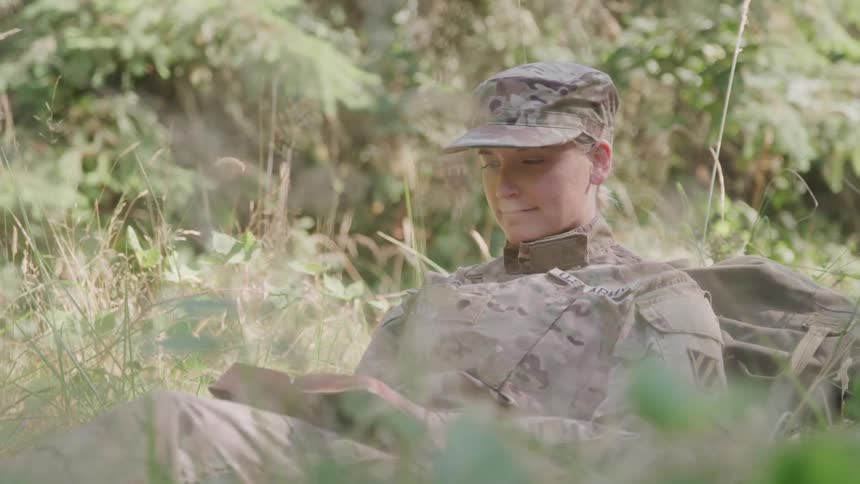 A military woman wearing her uniform and a hat lies on the grass in the jungle around midday reading a book with a smile on her face