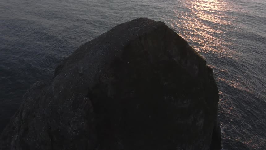 A large rocky cliff rises from the ocean with sunlight reflecting on the water
