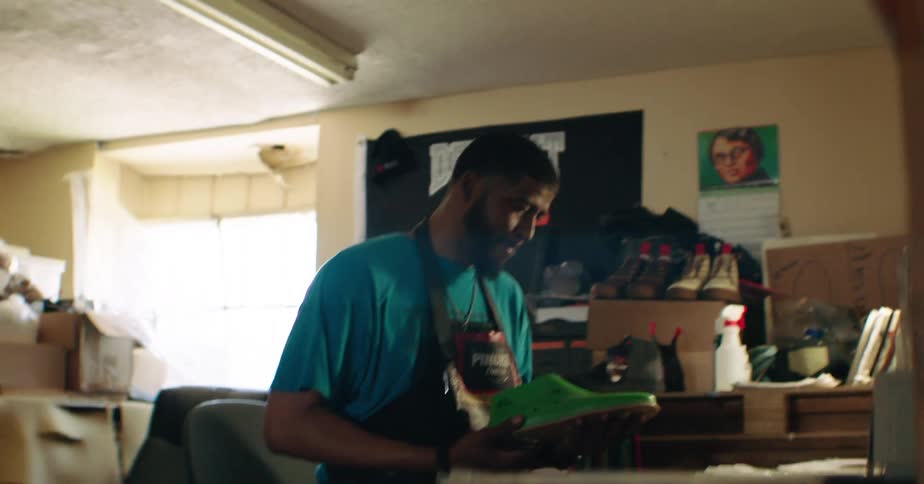 A man works attentively on a green shoe mold in a cluttered workshop