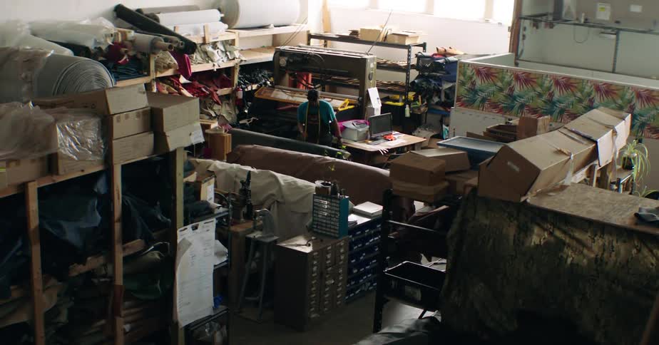 A person works in a cluttered industrial workshop surrounded by shelves materials and equipment