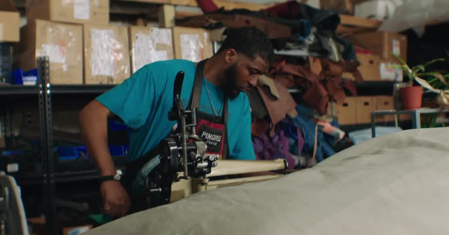 A man operates a sewing machine in a workshop crowded with fabric piles cardboard boxes and a potted plant