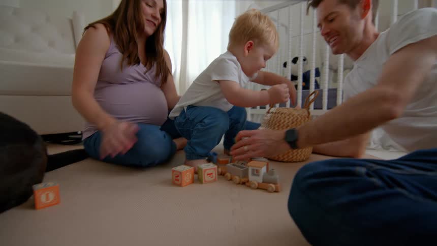 Pregnant mother playing with her little son next to father in a room with crib