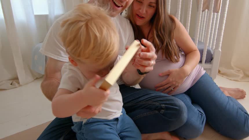 A child sits on her fathers lap clutching a framed photo while her pregnant mother smiles beside them in a warmly lit living room