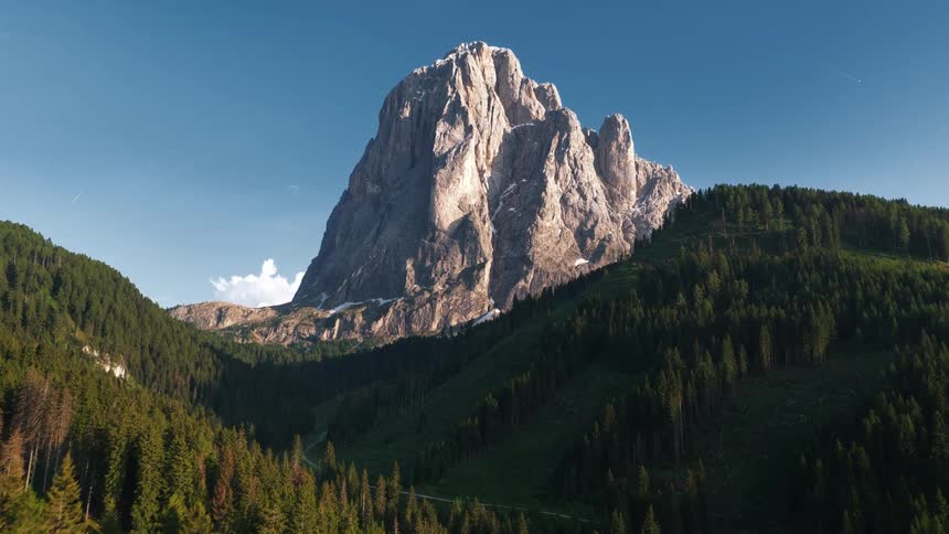 A towering rugged mountain peak rises sharply above a densely forested valley under a cloudless blue sky