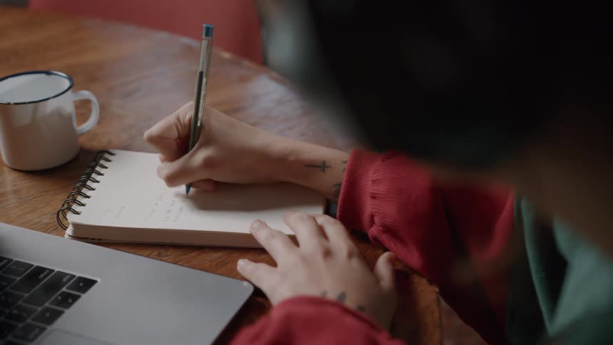 A person writes in a notebook beside a laptop and coffee mug on a wooden table