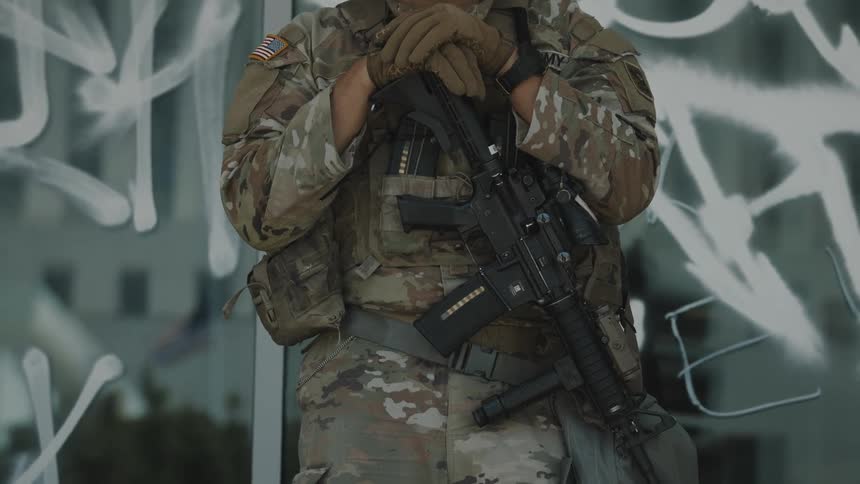 A national guard soldier in camouflage uniform stands guard holding a rifle in front of a graffiti covered window