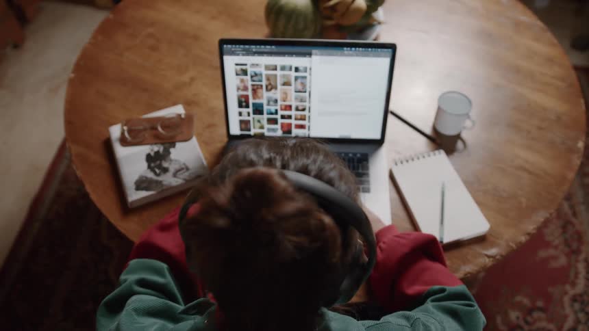 A person works on a laptop at a round wooden table with a notebook book and mug nearby