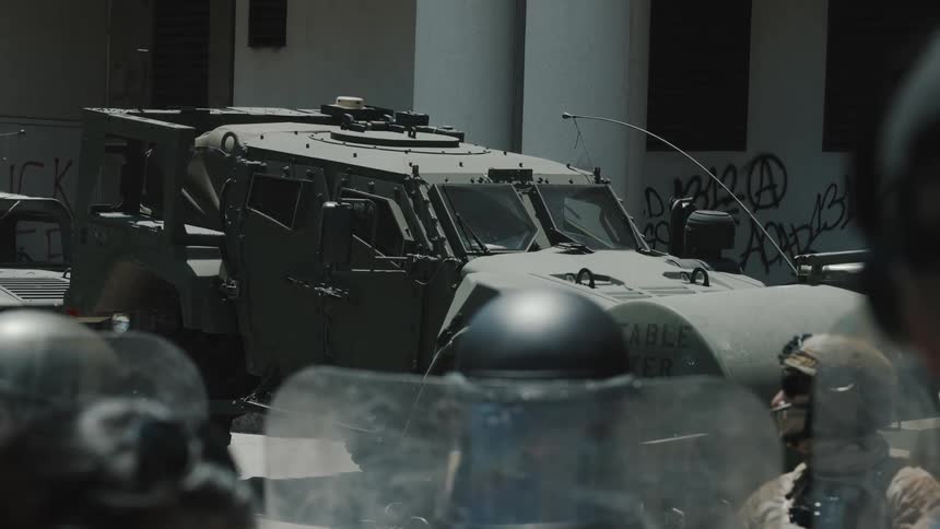 Riot police stand guard in front of a parked armored military vehicle during a tense standoff in an urban area with graffiti on the walls