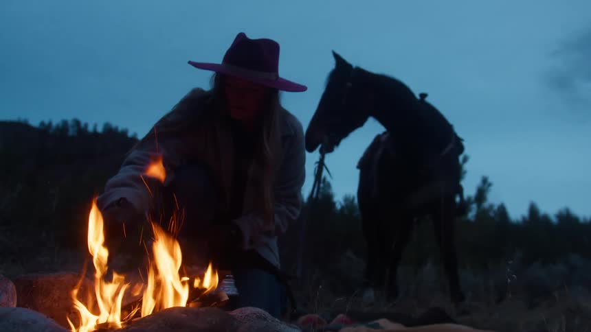 A person wearing a hat tends a small campfire beside a saddled horse in a twilight forest clearing