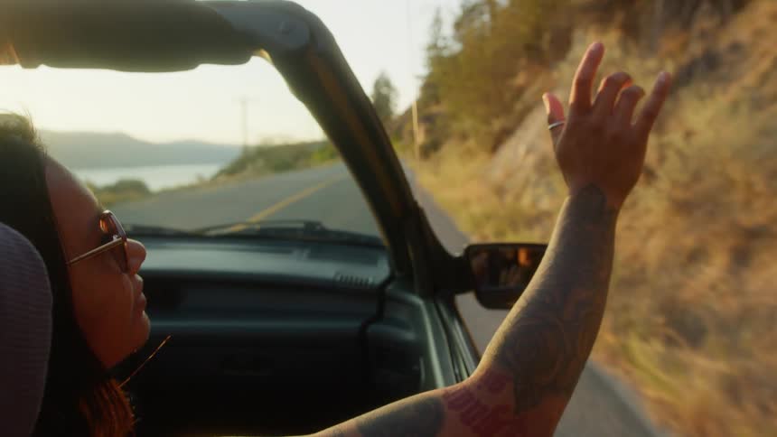 A young woman with a tattooed arm wearing sunglasses enjoys a joy ride in an openroof car sitting in the passenger seat she stretches her hands outside the car as it drives along a coastal road around midday
