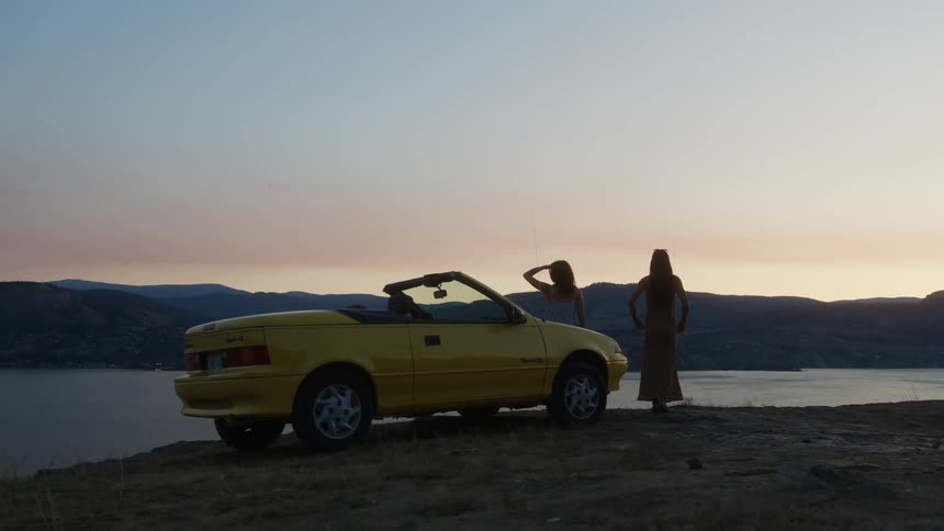 Two women stand beside a yellow convertible gazing at the sunset over a lake from a cliffside