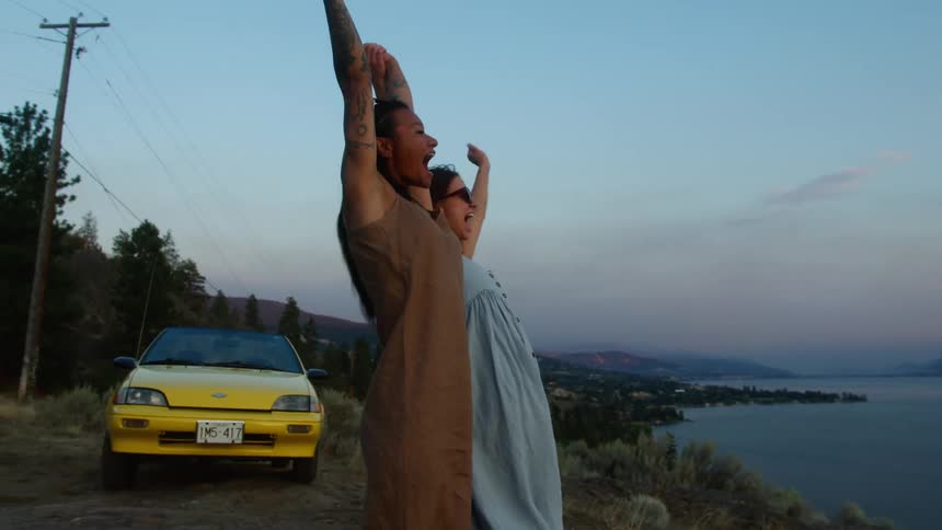 Two females standing near a lake at sunset shouts with their hands in the air