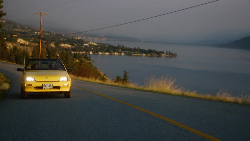 Two people inside a yellow convertible vehicle traveling along a seaside road