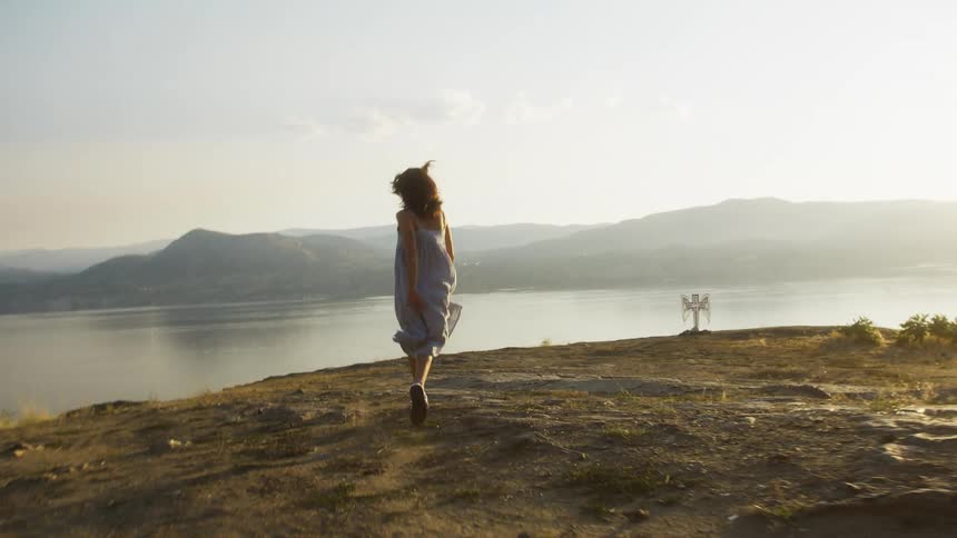 Woman in a flowing dress runs joyfully on a scenic hilltop at sunset near a large lake