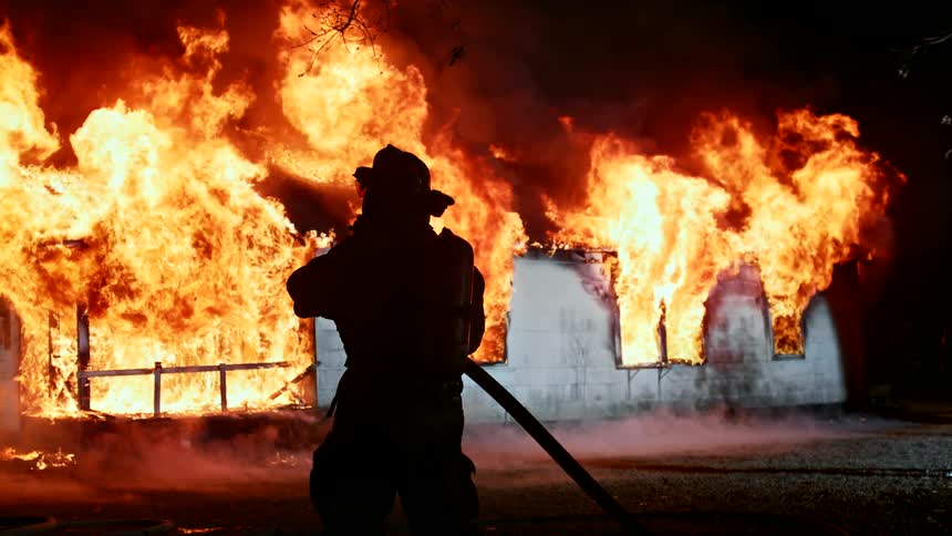 Firefighter stands in front of a burning building engulfed in a blaze of fire holding his hose spraying water